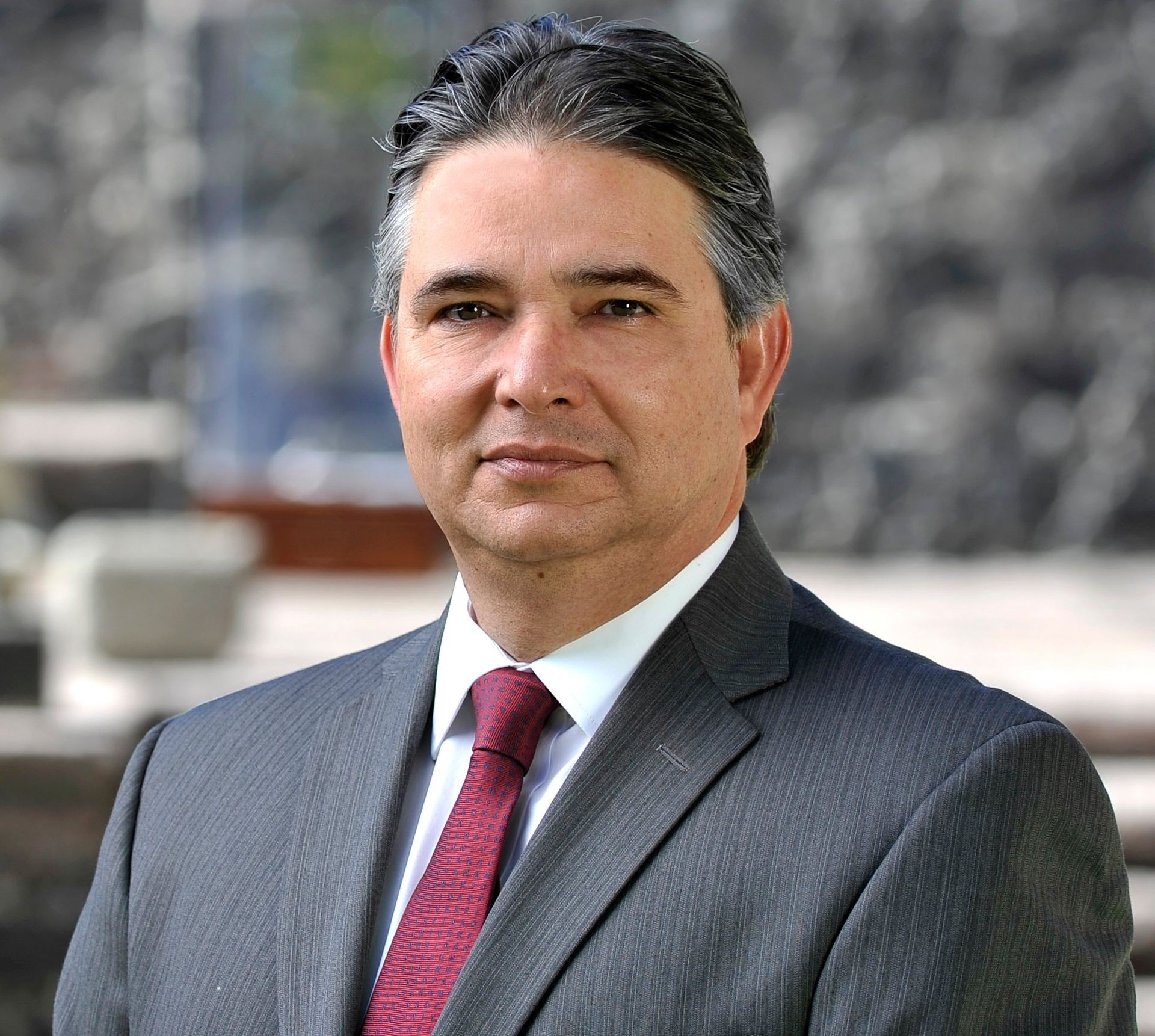 Man in suit and red tie standing in an outdoor setting with blurred background.