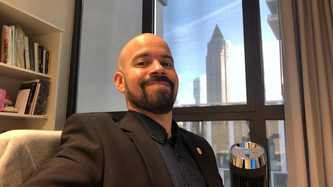 Person smiling in an office with a view of a cityscape through a window.