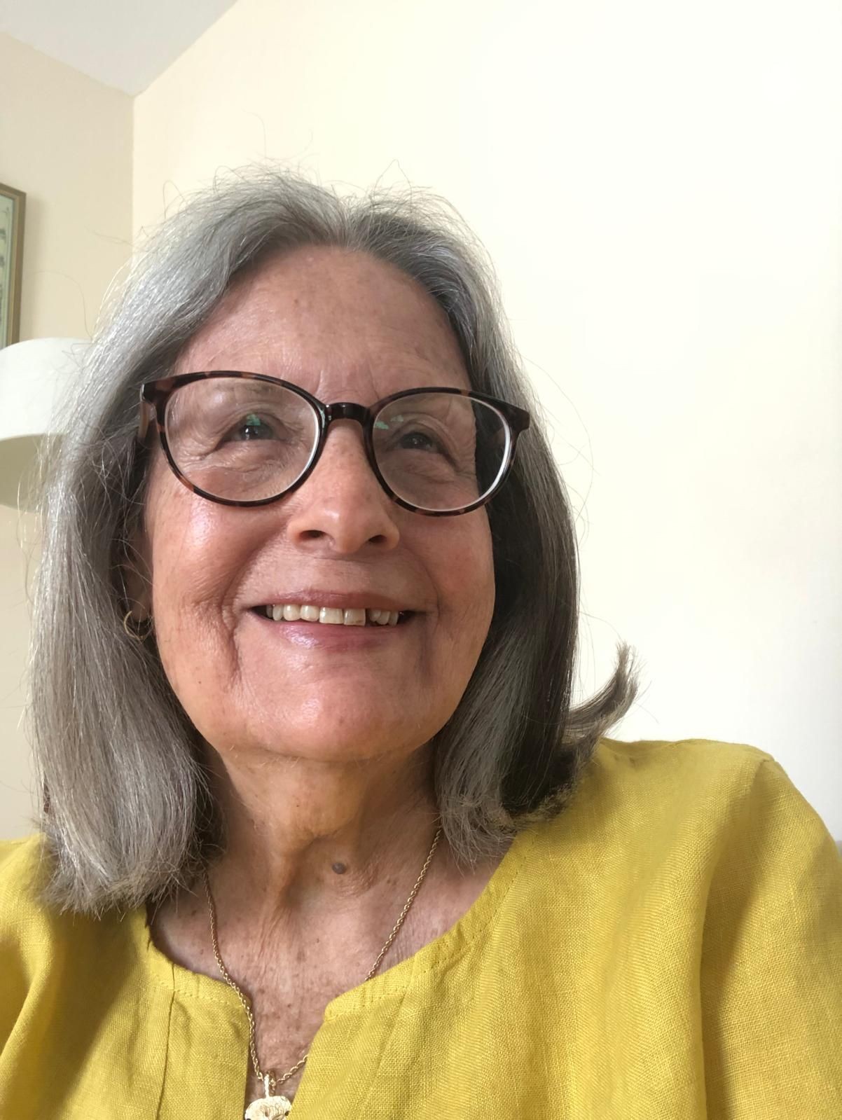 Person with shoulder-length grey hair wearing a yellow top and a gold necklace, sitting indoors.